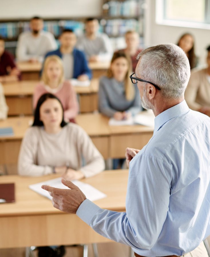 Back view of mature professor giving lecture to large group of college students in the classroom.